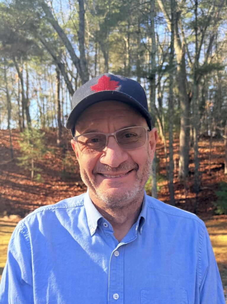 Selfie of a smiling man wearing a dark cap with a red maple leaf and a light blue shirt, outdoors among trees in autumn foliage.